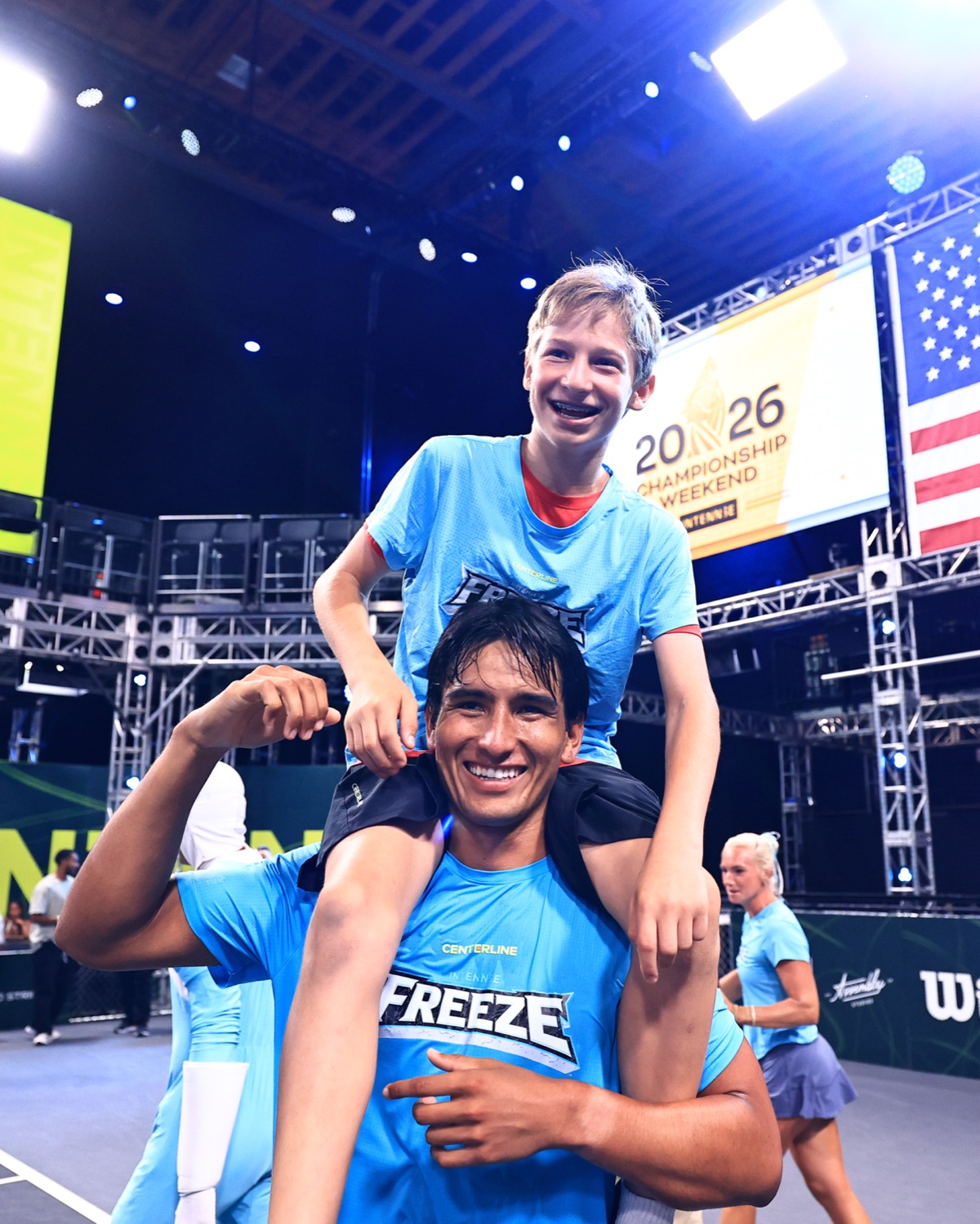 A young ball keeper riding on the shoulders of a player, both grinning, under Championship Weekend signage.