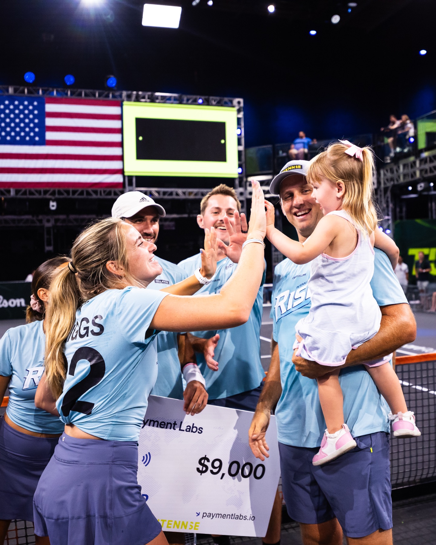 Players celebrating on court at a prize presentation, one lifting a child on their shoulders.
